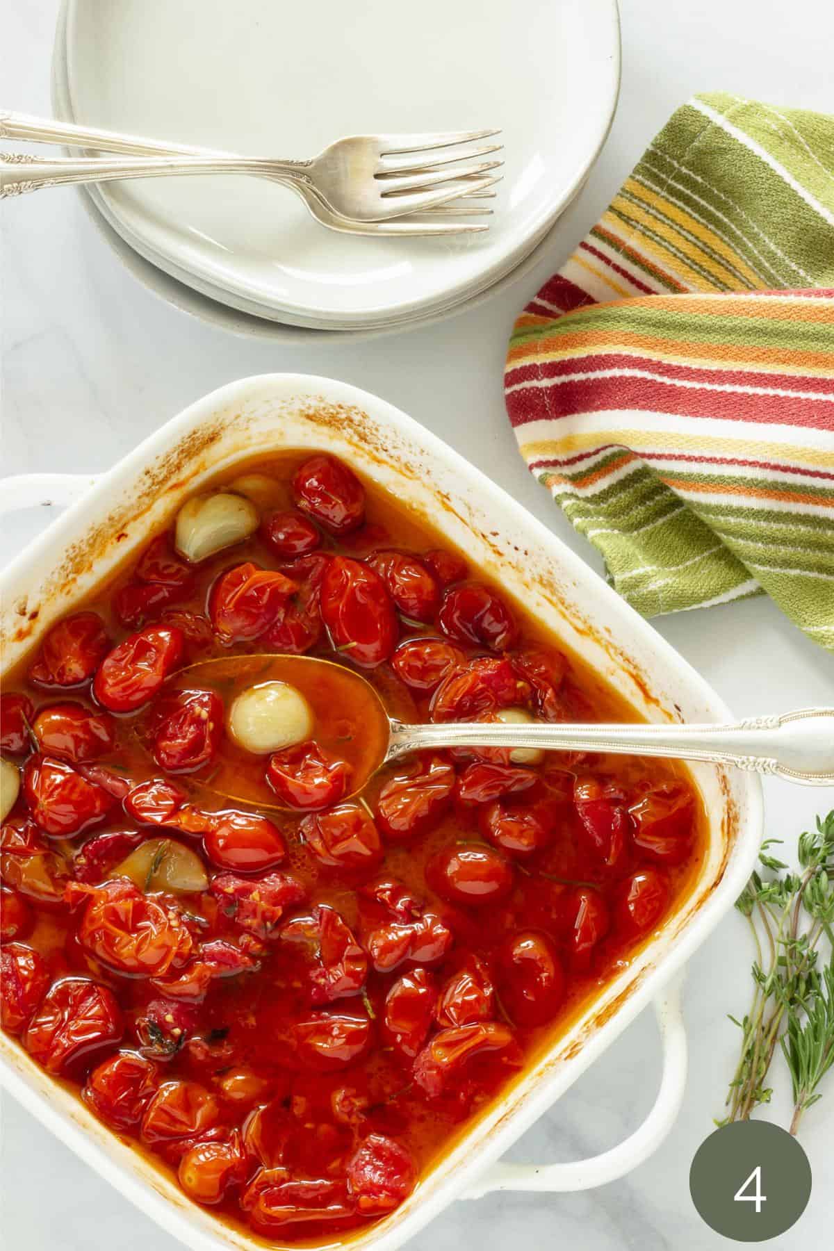 A finished dish of roasted tomatoes and garlic in a white casserole dish with a serving spoon 