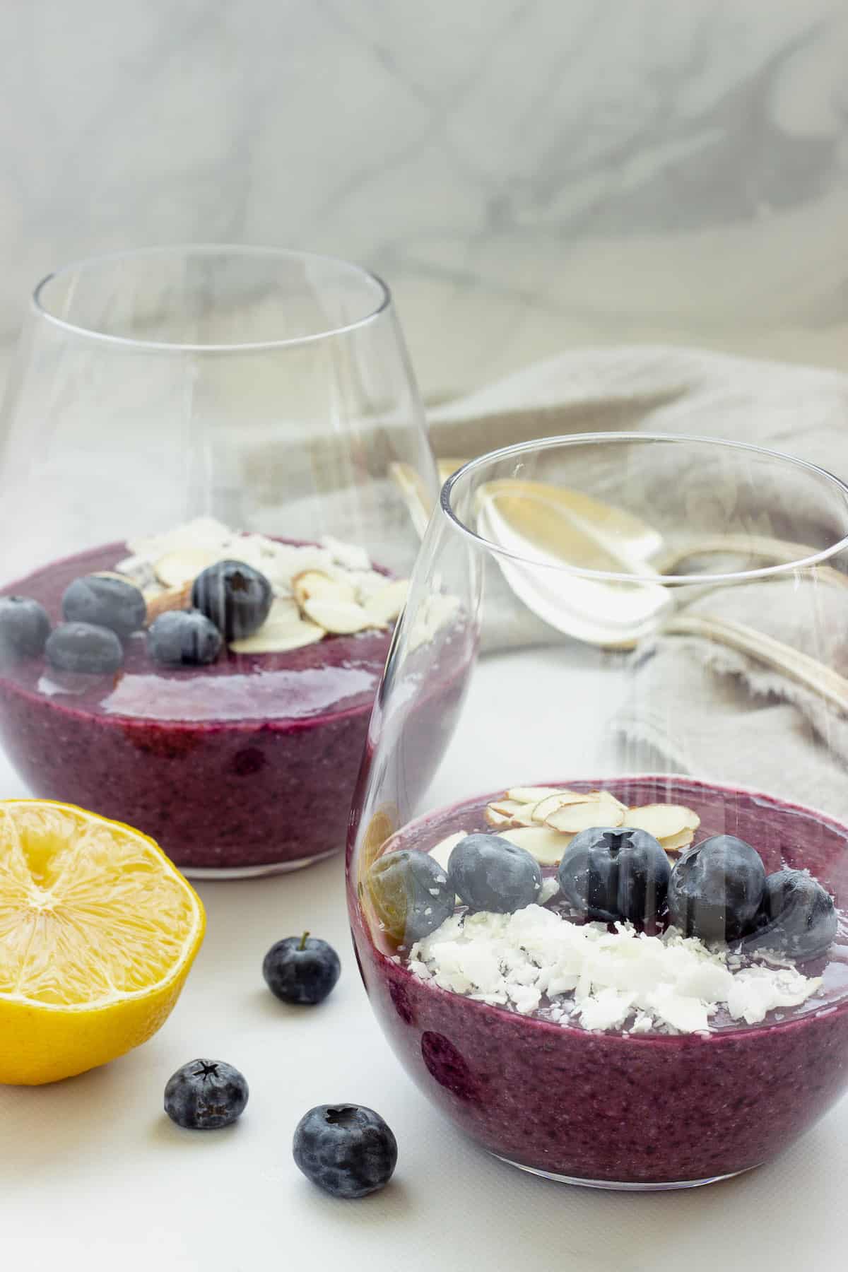 2 clear glasses with blueberry chia seed pudding garnished with fresh blueberries, coconut, and sliced almonds. A slice of lemon in the foreground and a spoon in the background.