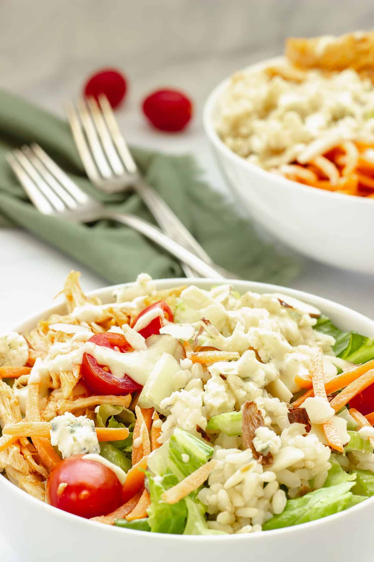 A white bowl full of Buffalo Chicken and Rice with vegetables and blue cheese dressing. A second bowl, green napkin, and two forks are in the background.