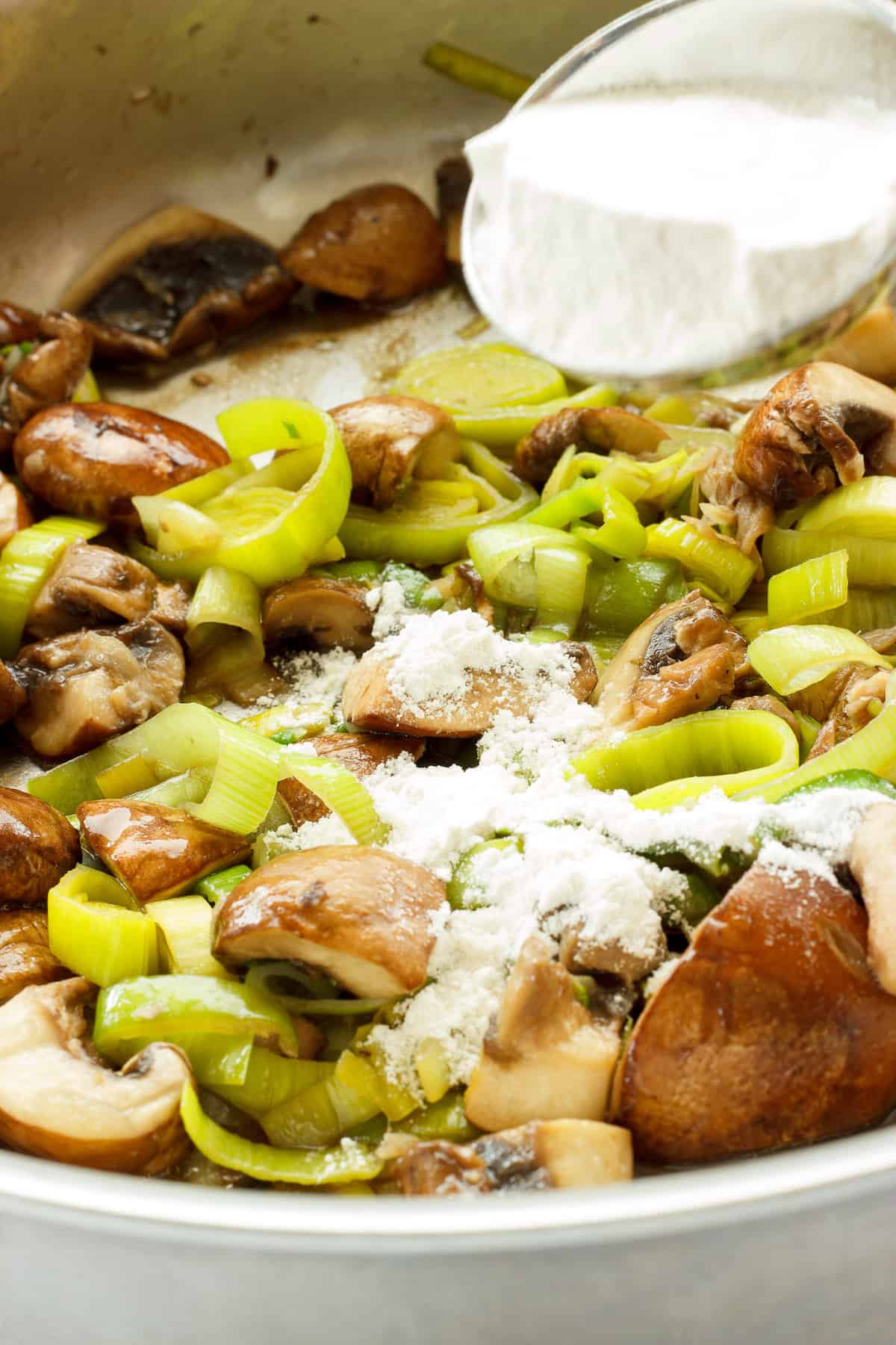 Instructions to make chicken with spinach and mushrooms. A spoon of flour sprinkled over cooked leeks and mushrooms in a saute pan.