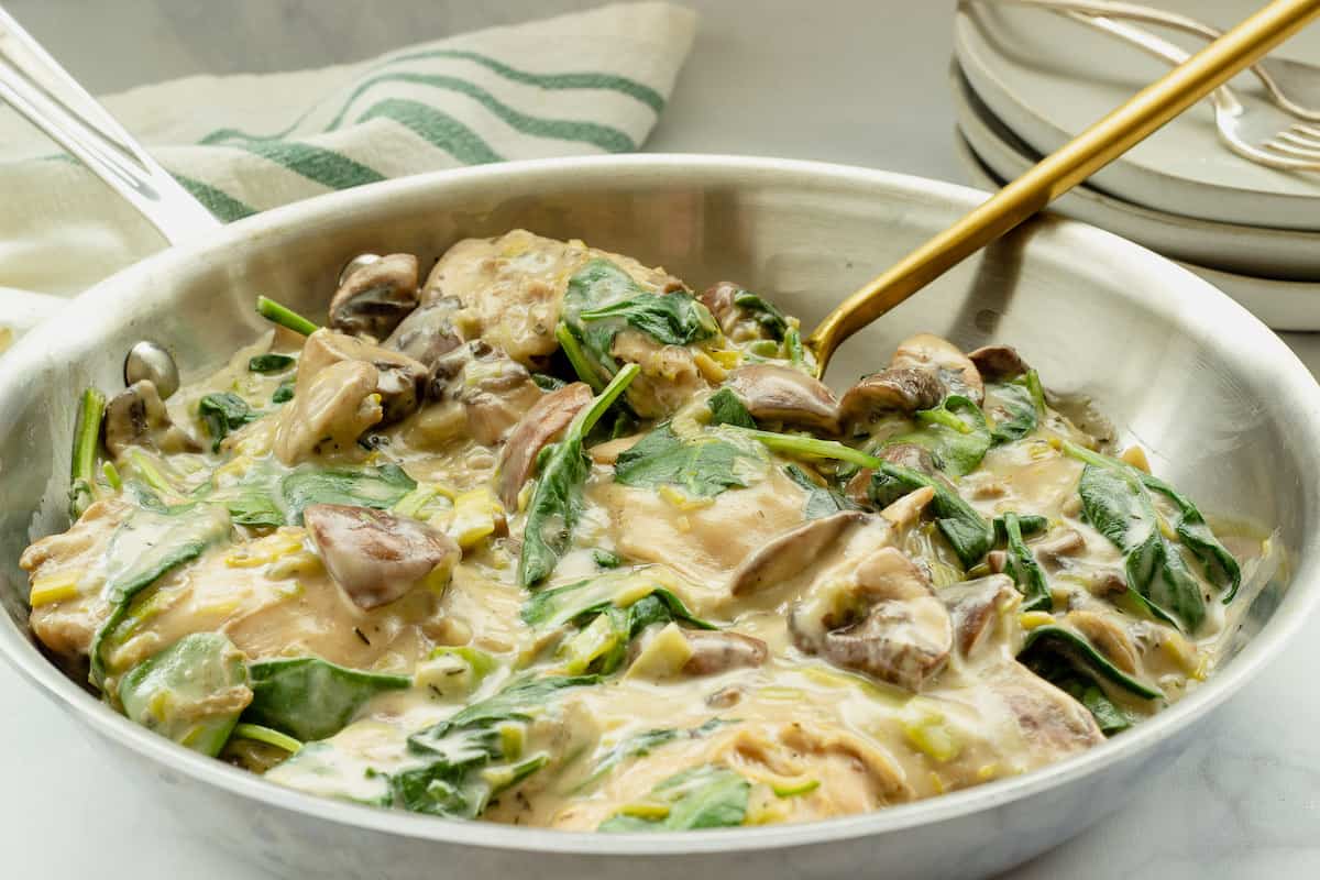 A saute pan of creamy chicken with spinach and mushrooms. A serving spoon is in the pan.