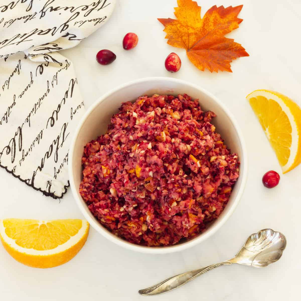A white bowl of cranberry orange relish. A serving spoon, napkin, fresh cranberries, orange slices and a fall leaf are in the background.