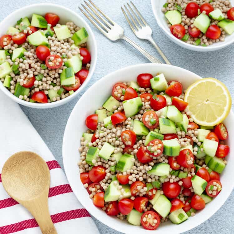 A white serving bowl and 2 smaller bowls of Cucumber Tomato Salad. A red and white striped napkin and wooden spoon are in the background.