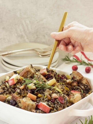 A person holding a gold spoon scooping wild rice stuffing casserole from a white casserole dish