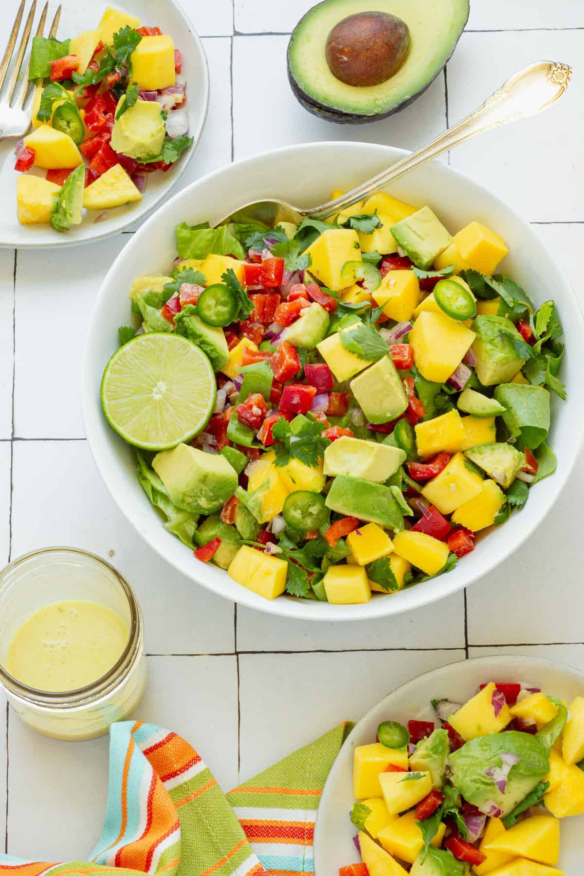 A white serving bowl with mango avocado salad. Two small serving plates with the salad, a jar of dressing, and a cut avocado in the background.