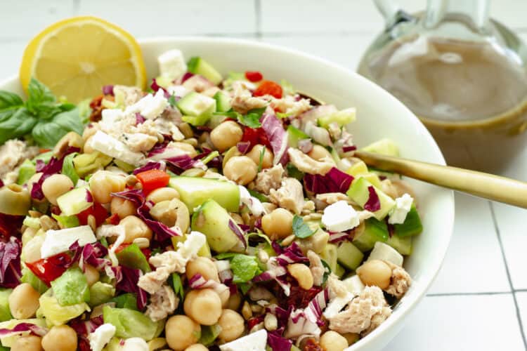 A white bowl of Mediterranean chopped salad. Serving plates, forks, and a bottle of dressing in the background.