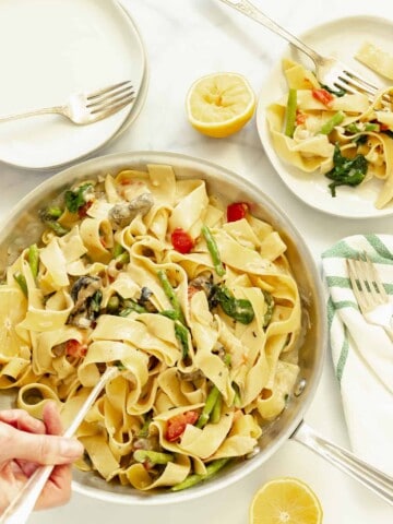 an overhead shot of a hand holding a serving spoon in a pan of pappardelle pasta primavera