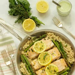 A pan of salmon fillets with rice and asparagus. Lemon halves and a bunch of herbs in the background