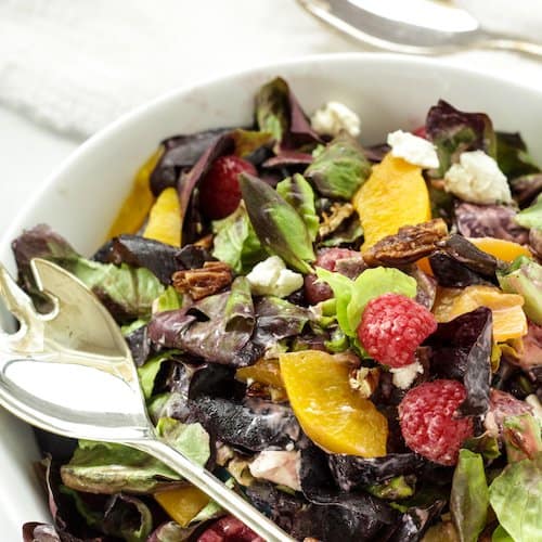 A closeup shot of a leafy green salad with peaches, raspberries, and goat cheese in a white bowl with serving spoons.