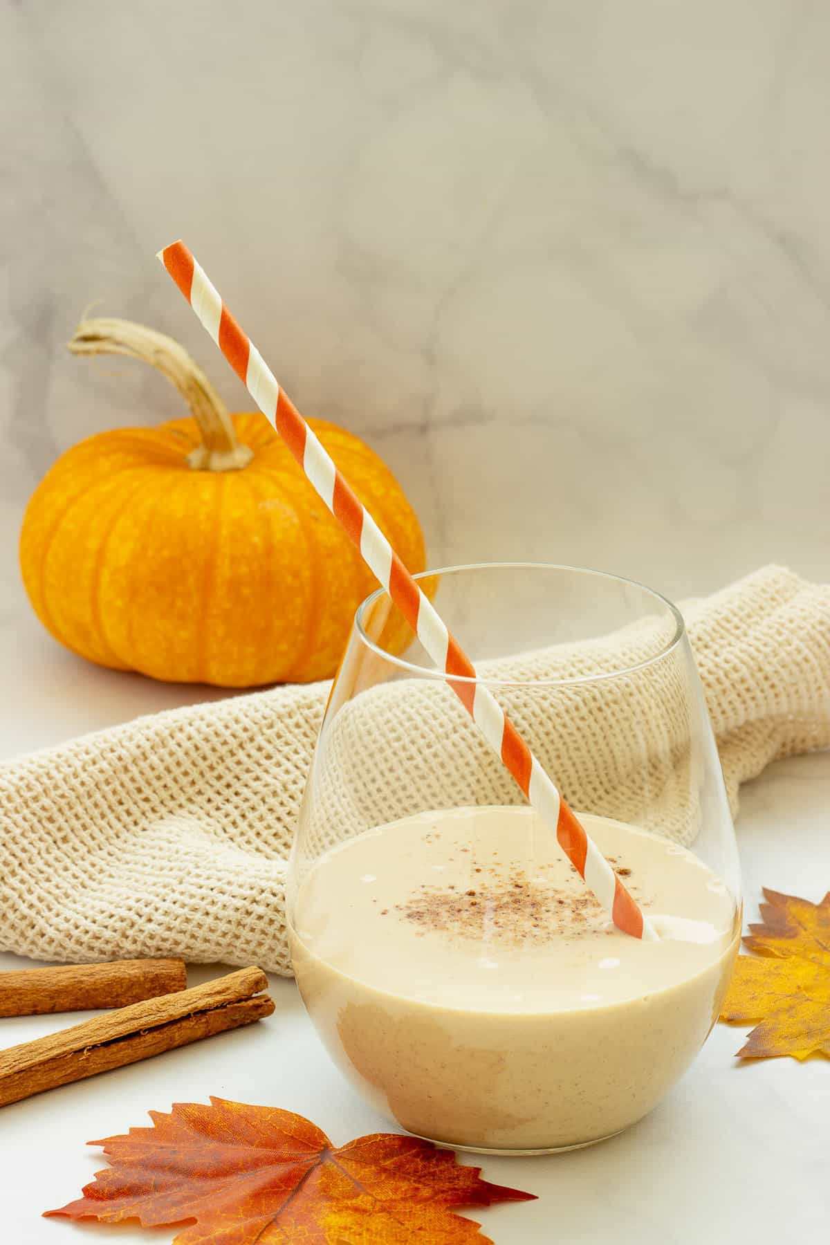 A glass of pumpkin pie smoothie with an orange and white striped straw. Fall leaves and a mini pumpkin are in the background.