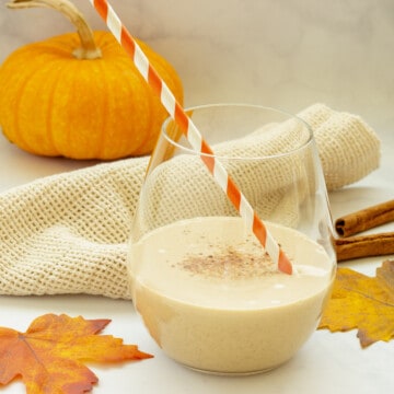 A glass of pumpkin pie smoothie with an orange and white striped straw. Fall leaves and a mini pumpkin are in the background.