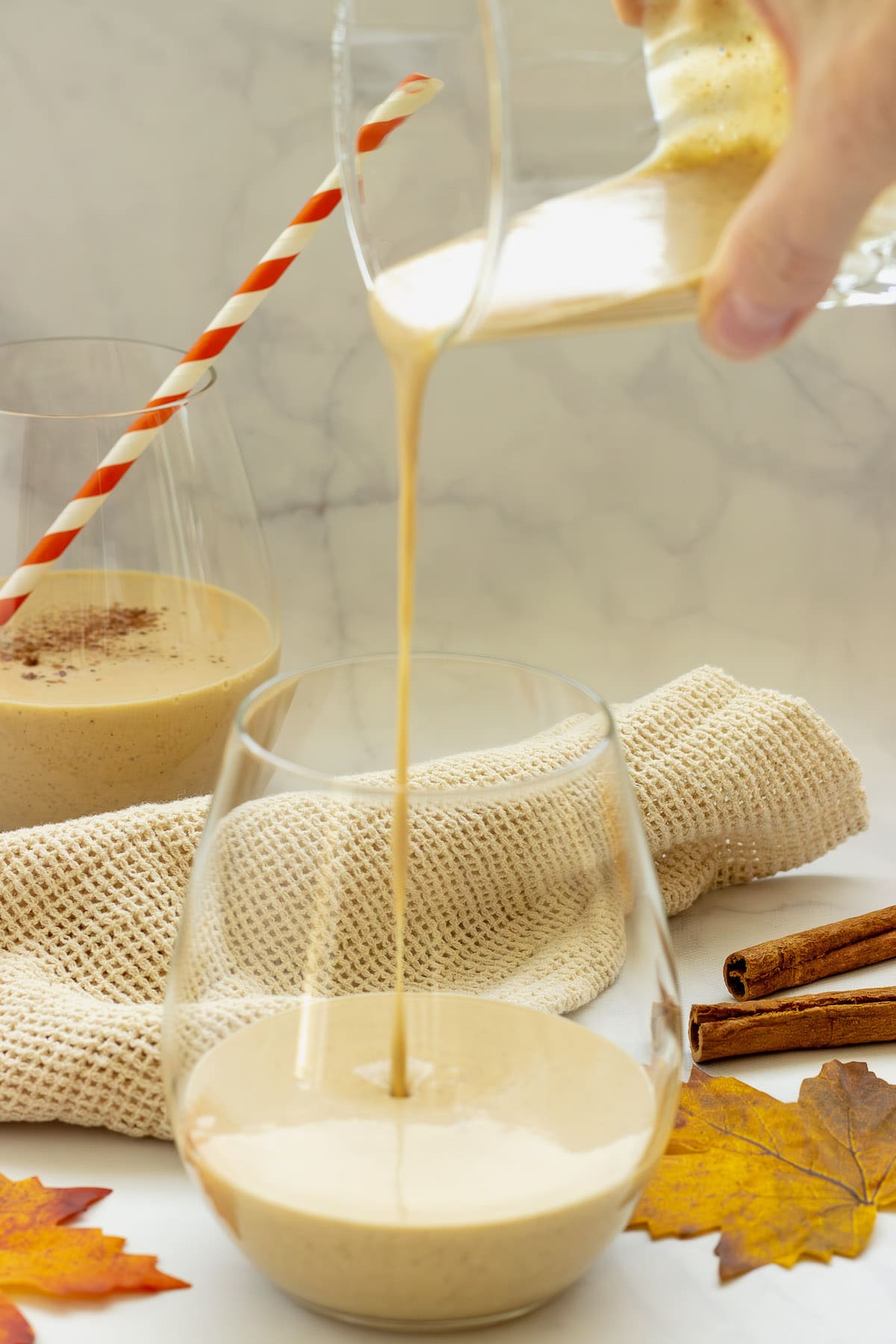 A person pouring pumpkin pie smoothie into a glass. Another glass of pumpkin pie smoothie with a straw is in the background.