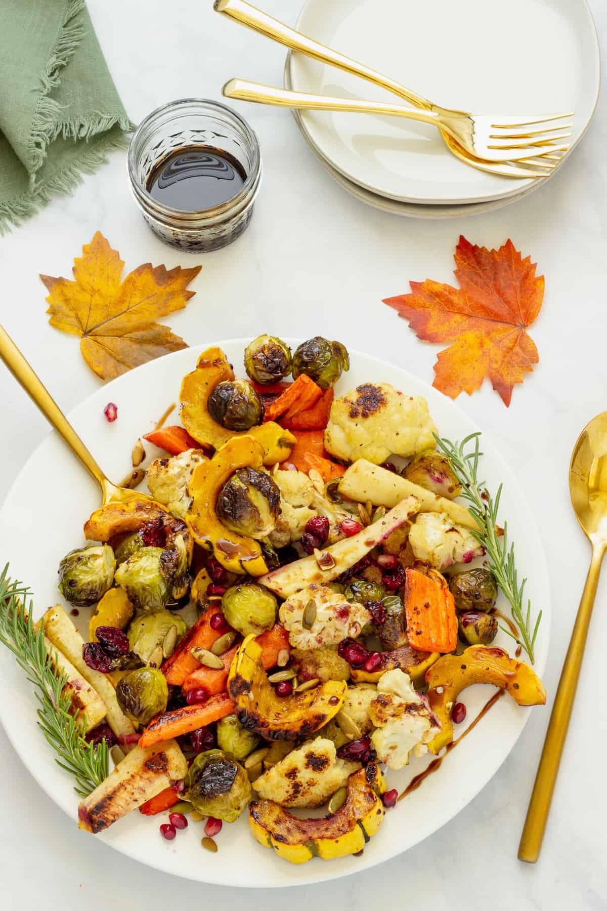 A white platter of roasted fall vegetables with a small jar of balsamic glaze. Fall leaves, serving spoons, forks, and plates are in the background.
