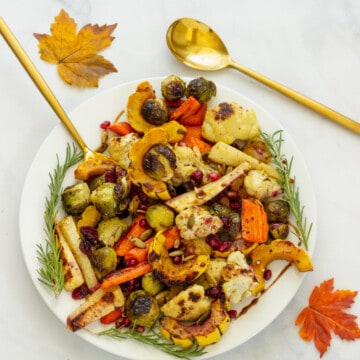 A white platter of roasted fall vegetables with fall leaves and gold serving spoons in the background.