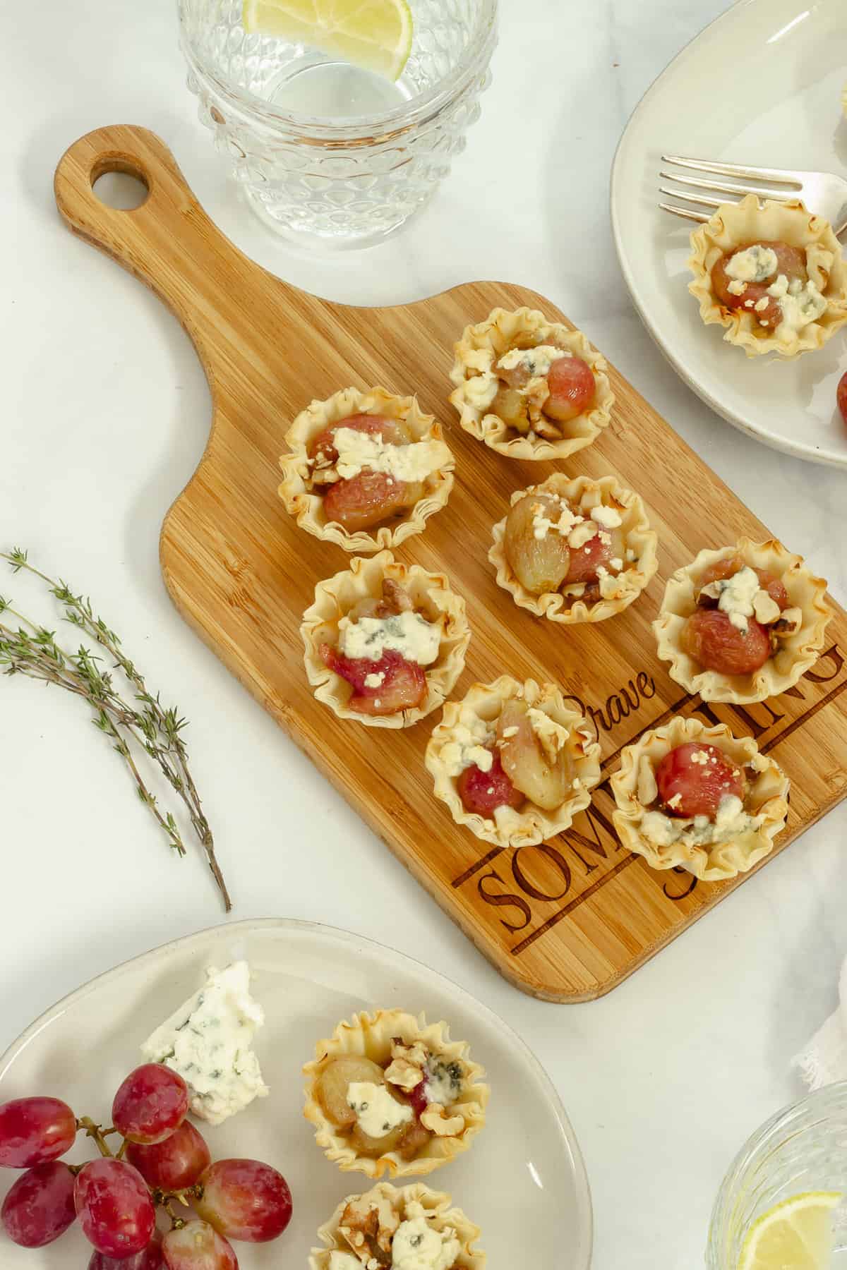 A cheese board with roasted grapes phyllo cup appetizers. Two serving plates with cheese, grapes, and the appetizer cups are in the background.