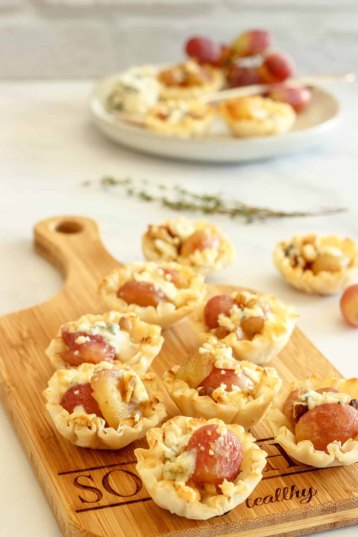 A serving board with roasted grapes phyllo cup appetizers. A plate of appetizers is in the background.
