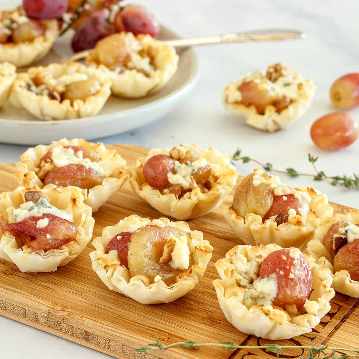 A serving board with roasted grapes phyllo cup appetizers. A plate of appetizers with fresh grapes is in the background.