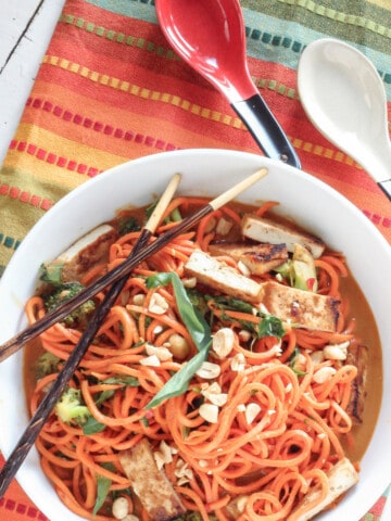 A white bowl of sesame peanut sweet potato noodles with spoons and a striped napkin in the background.