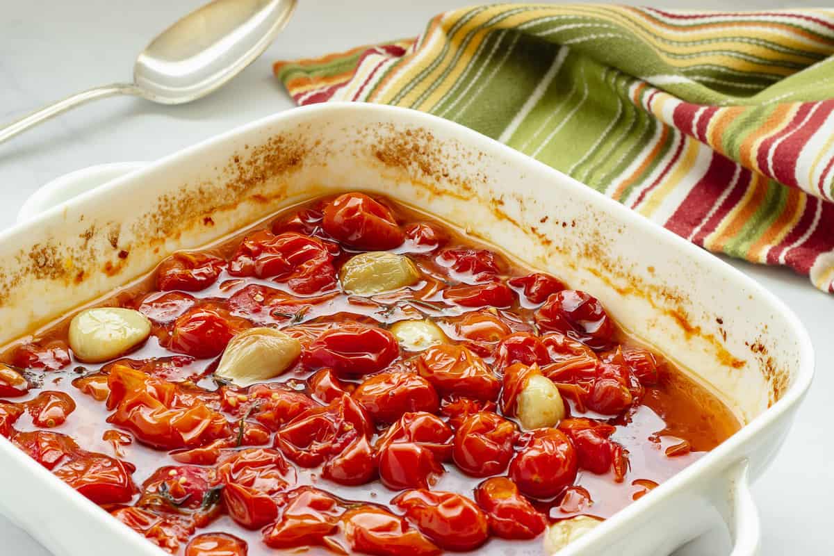 A baking dish with slow roasted grape tomatoes and garlic in olive oil. A striped napkin is in the background.