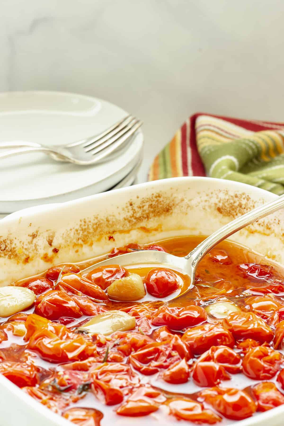 A spoon scooping slow roasted grape tomatoes out of a casserole baking dish. A stack of white plates with forks and a striped napkin in the background.