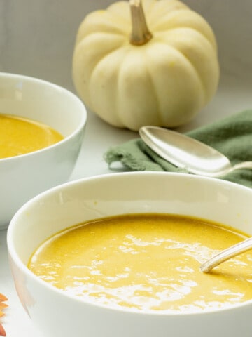 Two white bowls of spiced pumpkin bisque soup with a green napkin and white pumpkin in the background.