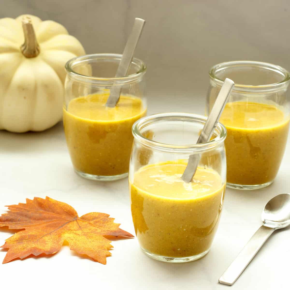 Three small appetizer glasses filled with spiced pumpkin bisque. Mini spoons, a fall leaf, and a white pumpkin are in the background.