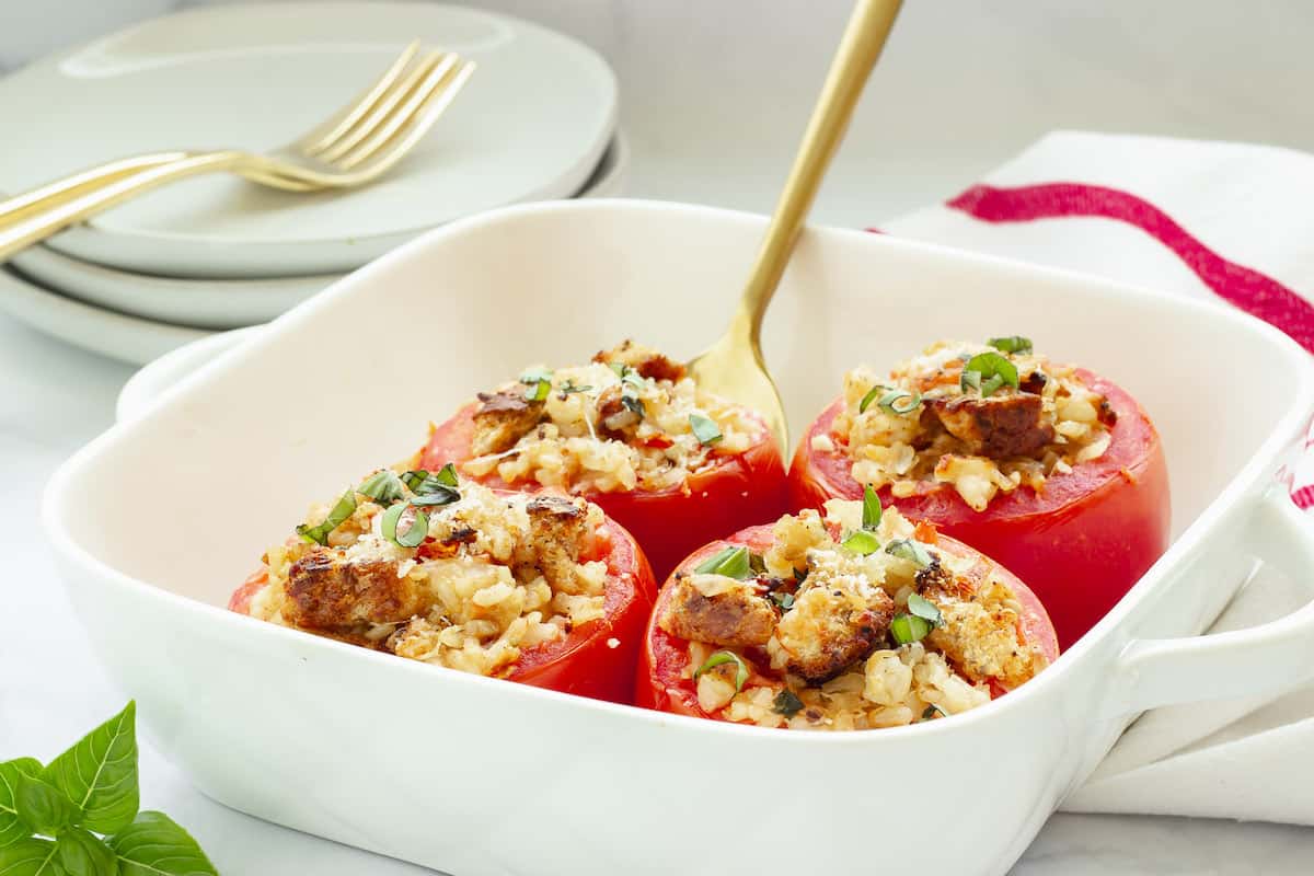 A white casserole dish with 4 baked stuffed tomatoes and a gold serving spoon under one tomato. Plates, forks, and a red and white striped napkin in the background.