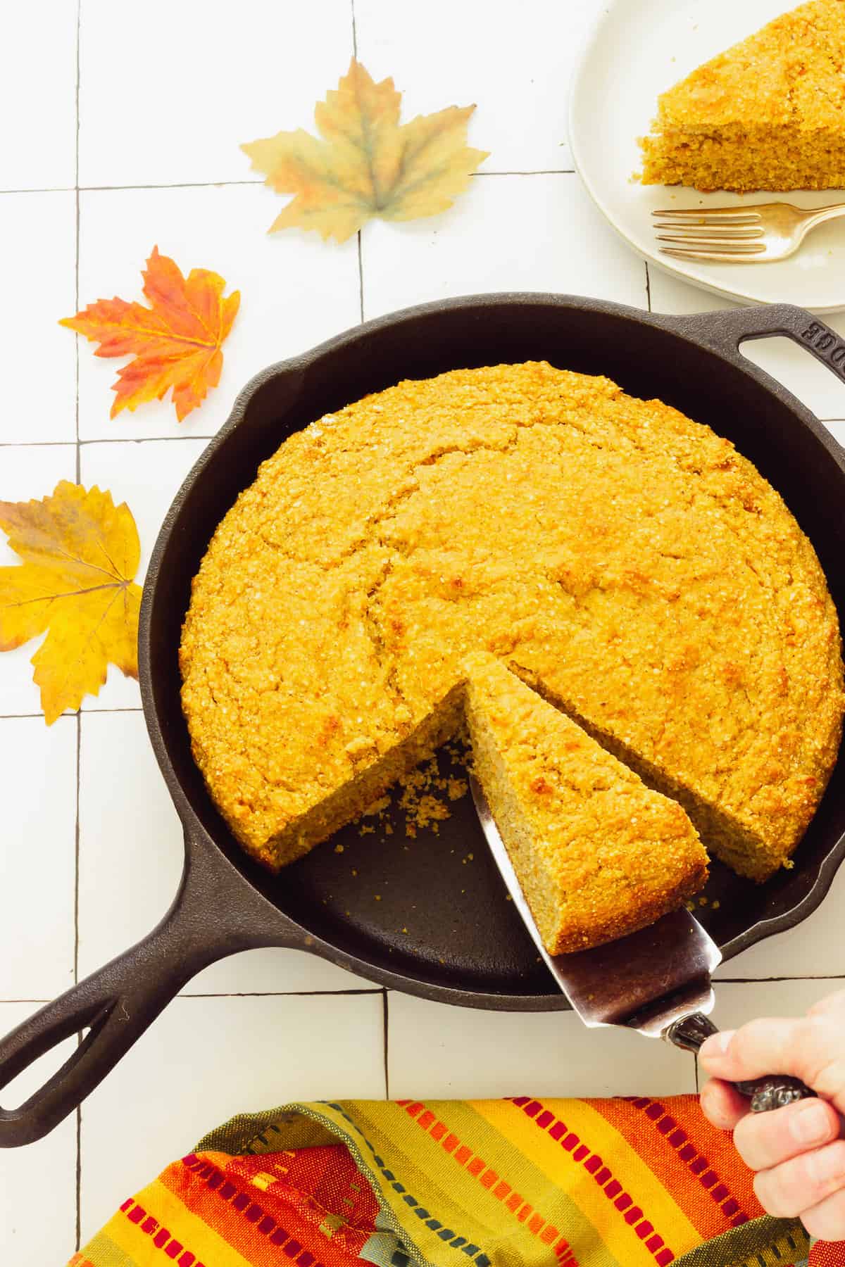 A cast iron skillet with sweet potato cornbread. Fall leaves, a fall napkin, and a small plate with a slice of cornbread are in the background.