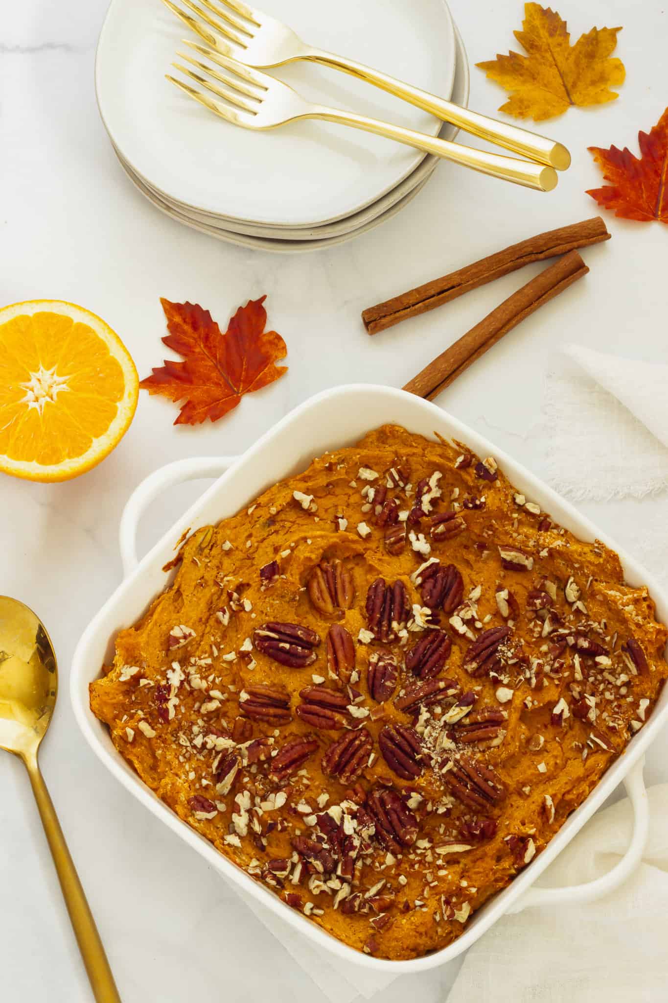 A square white casserole dish with sweet potato souffle. Serving plates, utensils, fall leaves, and cinnamon sticks are in the background.