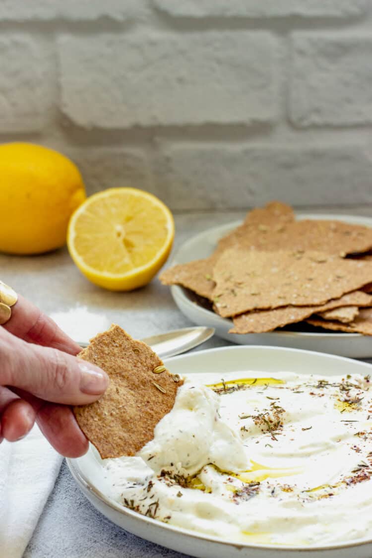 A person's hand with a cracker dipping into a plate of whipped feta dip. Lemons and a plate of crackers are in the background.